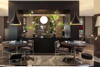 Dining area with round black tables, gray chairs, plants in transparent vases, and hanging black pendant lights at B&B Hotel Arezzo
