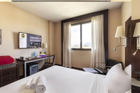 Hotel room at B&B Hotel Arezzo with a bed, rolled towels, a desk with a laptop, TV on the wall, and a window with curtains