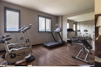 Gym room at B&B Hotel Arezzo with exercise bikes, treadmill, dumbbells, wall mirror, two windows, and a water dispenser