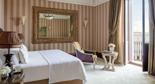 Bedroom with white bed linens, large ornate mirror, armchair, small round table with flowers, and balcony view at Anantara Palazzo Naiadi Rome Anantara Palazz
