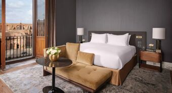 Hotel room at Anantara Palazzo Naiadi Rome Hotel Anantara Palazz featuring a bed with white linens, brown sofa, table with flower vase, and balcony view of historical buildings