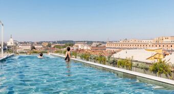 Two people swimming and standing in a rooftop pool overlooking city buildings under a clear blue sky at Anantara Palazzo Naiadi Rome Hotel Anantara Palazz