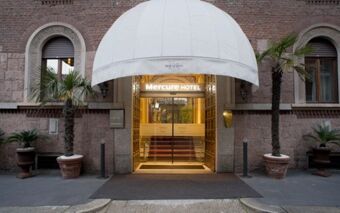 Entrance of Hotel Regency Milano with white dome canopy, glass doors, brown brick walls, and two tall potted palm trees