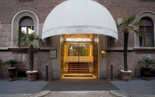 Entrance of Hotel Regency Milano with white dome canopy, glass doors, brown brick walls, and two tall potted palm trees