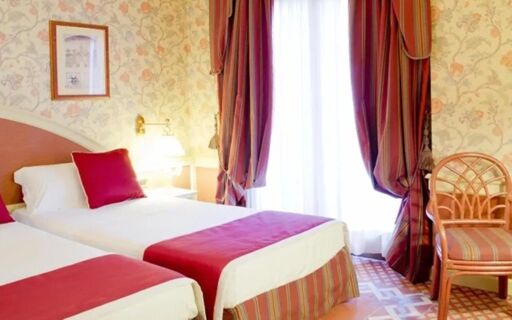 Two single beds with red pillows and blankets in a floral wallpaper room at Hotel Regency Milano with a window and chair