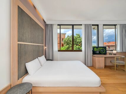 Modern hotel room at B&B Hotel Padova Methis with a large bed, wooden headboard, windows with curtains, and a desk with a TV and chair