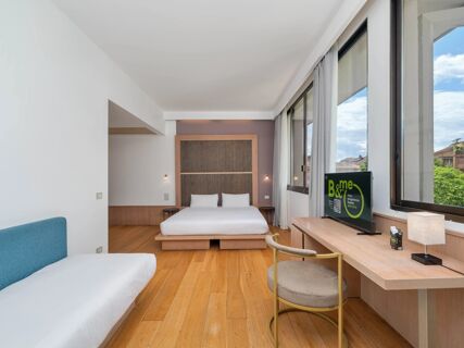 Hotel room at B&B Hotel Padova Methis with a bed, blue sofa, wooden flooring, desk with chair, and large windows with curtains