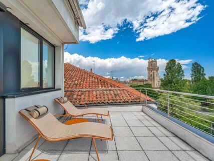 Two beige lounge chairs with cylindrical pillows on a rooftop terrace with a red tile roof and a tall tower in the distance at B&B Hotel Padova Methis