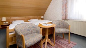 Double bed with white linens under a wooden sloped ceiling, two patterned armchairs and a round wooden table at Gasthof Zur Linde
