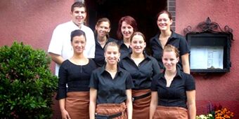 Seven Gasthof Zur Linde staff members wearing black shirts and brown aprons stand in front of a red building wall with a menu display case