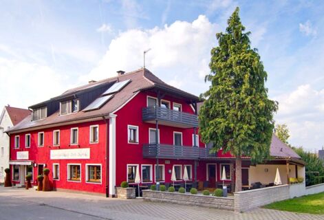Red building with balconies, outdoor seating, a large tree, and signs reading Gasthof Zur Linde under a partly cloudy sky
