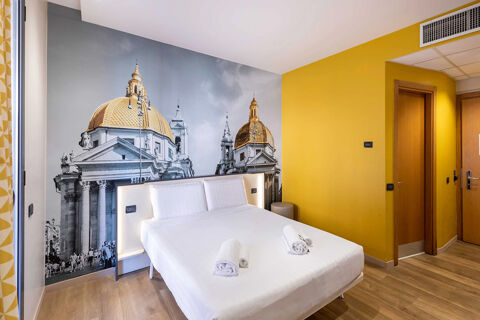 Modern hotel room with a large bed, rolled white towels, yellow walls, wood flooring, and a cityscape mural at B&B Hotel Roma Tuscolana San Giovanni