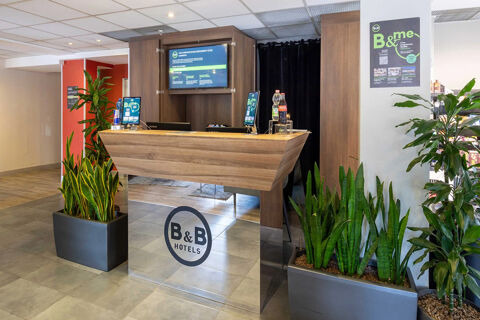 Reception desk with B&B Hotels logo, two plants in large gray pots, digital information screens, and bottled drinks at B&B Hotel Roma Tuscolana San Giovanni