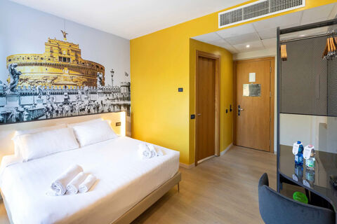 Hotel room at B&B Hotel Roma Tuscolana San Giovanni with a large bed, yellow wall, wooden doors, desk, and city mural above the bed