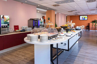 Buffet style breakfast setup with plates, covered cold cuts, fruits, pastries, and beverage machines at B&B Hotel Roma Tuscolana San Giovanni