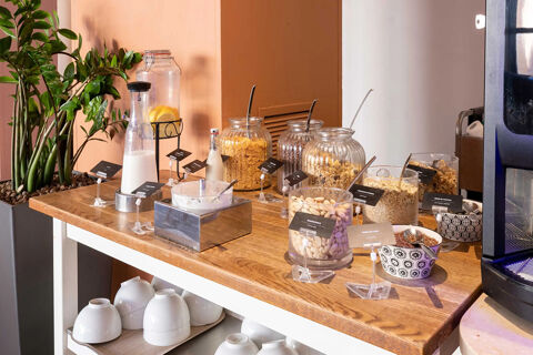 Buffet setup with jars of cereals, nuts, milk, bowls, and plants on a wooden table at B&B Hotel Roma Tuscolana San Giovanni