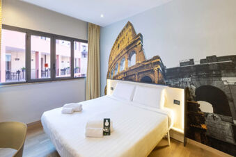 Hotel room with double bed, white linens, towels, large window, and a Colosseum mural at B&B Hotel Roma Tuscolana San Giovanni