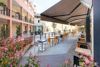 Outdoor seating area with black and orange chairs, tables, large sun shades, and pink flowers at B&B Hotel Roma Tuscolana San Giovanni