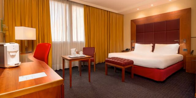 Hotel room at Crowne Plaza Venice East featuring a double bed with red headboard, wooden furniture, yellow curtains, and a coffee machine