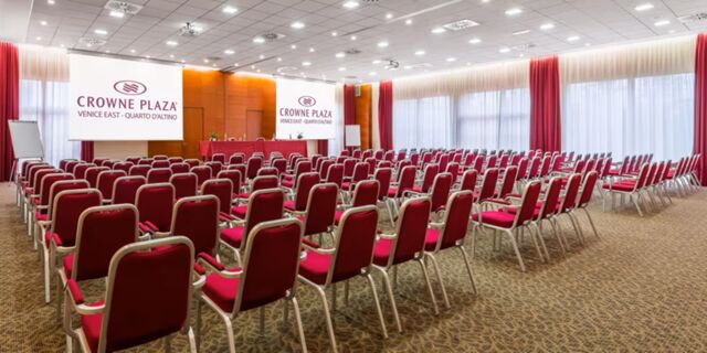 Conference room at Crowne Plaza Venice East with rows of red chairs facing two large projection screens and windows with red curtains