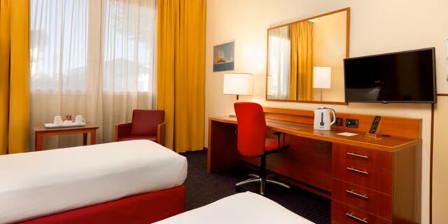 Hotel room at Crowne Plaza Venice East with two beds, a red chair, desk with kettle, wall-mounted TV, and yellow curtains