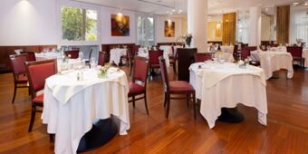 A dining area at Crowne Plaza Venice East with round tables covered in white cloths and red cushioned wooden chairs on a wooden floor