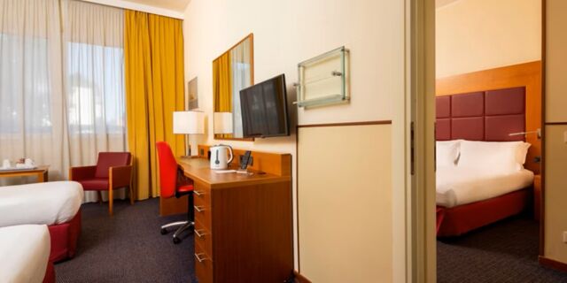 Hotel room at Crowne Plaza Venice East with two beds, a desk with TV, a red chair, and yellow curtains on the window