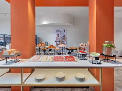 Buffet table with sliced bread, croissants, cold cuts, cheeses, fruits, and infused water at B&B Hotel Milano Cologno Studios
