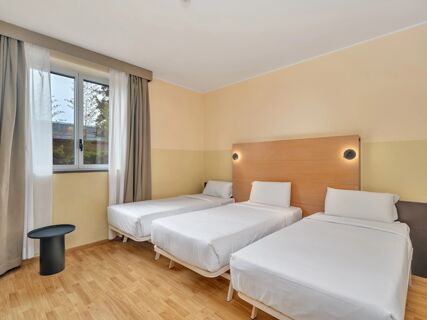 Three single beds with white linens and pillows are placed against a beige wall with modern lights at B&B Hotel Milano Cologno Studios