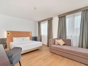 Bright hotel room at B&B Hotel Milano Cologno Studios with a double bed, sofa, desk, chair, and two windows with curtains