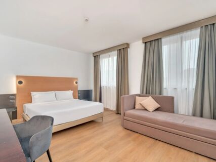 Bright hotel room at B&B Hotel Milano Cologno Studios with a double bed, sofa, desk, chair, and two windows with curtains