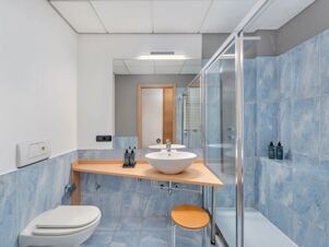 Bathroom with blue tiles, glass shower cabin, white sink on wooden counter, small stool, and mirror at B&B Hotel Milano Cologno