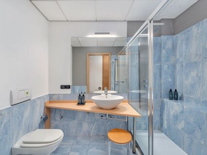 Bathroom with blue tiles, glass shower cabin, white sink on wooden counter, small stool, and mirror at B&B Hotel Milano Cologno