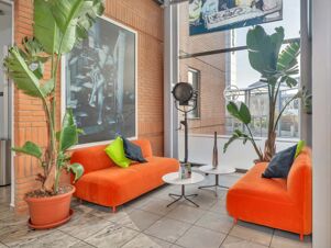 Two orange sofas with green and black pillows, potted tall plants, black vintage lamp, and black and white photos at B&B Hotel Milano Cologno Studios
