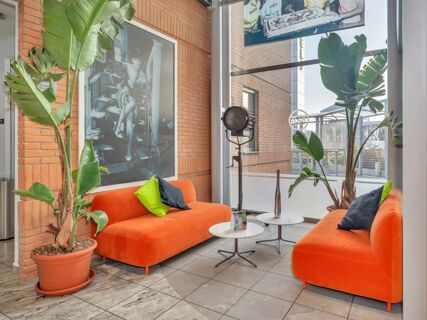 Two orange sofas with green and black pillows, potted tall plants, black vintage lamp, and black and white photos at B&B Hotel Milano Cologno Studios