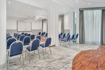 Conference room with rows of blue chairs, a projector screen, and large windows with curtains at B&B Hotel Milano Cologno Studios