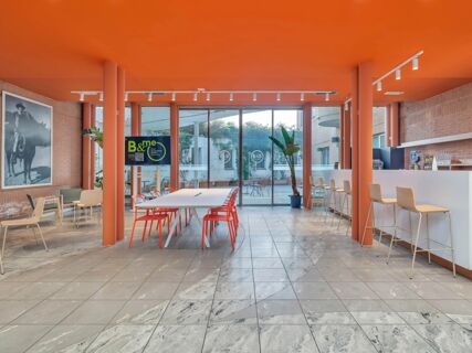 Interior of B&B Hotel Milano Cologno with orange ceiling and pillars, long white table with orange chairs, bar area, and glass doors leading outside