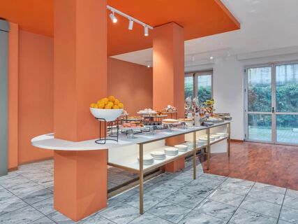 Breakfast buffet table with assorted pastries, fresh oranges, and plates in a room with orange walls at B&B Hotel Milano Cologno Studios