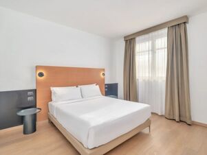 A neatly made bed with white linens and pillows, wooden headboard with two round wall lights, beige curtains and a small dark side table in a room at B&B Hotel Milano Cologno Studios
