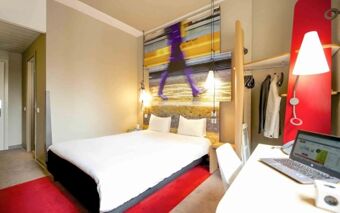 Hotel ibis Milano Centro room with a double bed, red carpet, a large wall photo, hanging clothes, and a desk with a laptop and lamp