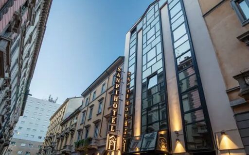 The exterior of Hotel Sanpi Milano is shown with illuminated signage on a multi-story building against a clear evening sky