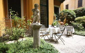 Stone cherub statue on a pedestal in a garden courtyard with a table, four chairs, plants, and Hotel Sanpi Milano walls