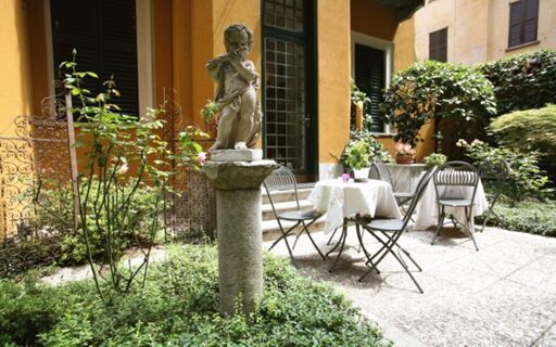Stone cherub statue on a pedestal in a garden courtyard with a table, four chairs, plants, and Hotel Sanpi Milano walls