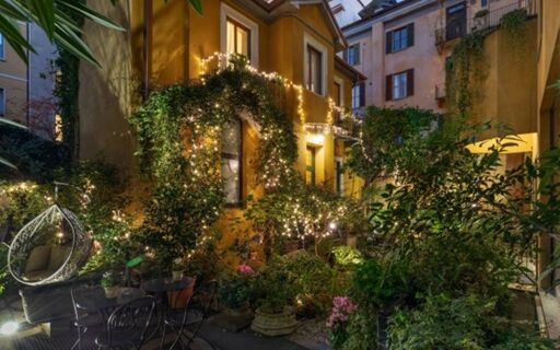 A nighttime view of Hotel Sanpi Milano's courtyard with illuminated plants, a hanging chair, tables, and lush greenery