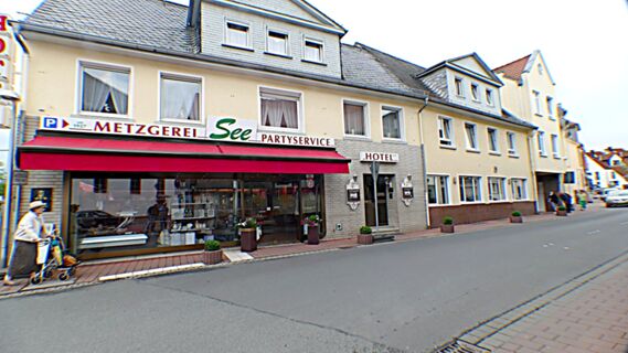Die Stra&szlig;enansicht von Hotel & Metzgerei See zeigt ein mehrst&ouml;ckiges Geb&auml;ude mit Metzgerei, Partyservice und Hoteleingang in einer ruhigen Ortschaft.