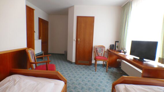 Helles G&auml;stezimmer im Hotel & Metzgerei See mit zwei Einzelbetten, zwei bunten St&uuml;hlen, Schreibtisch und Fenster mit Vorh&auml;ngen.