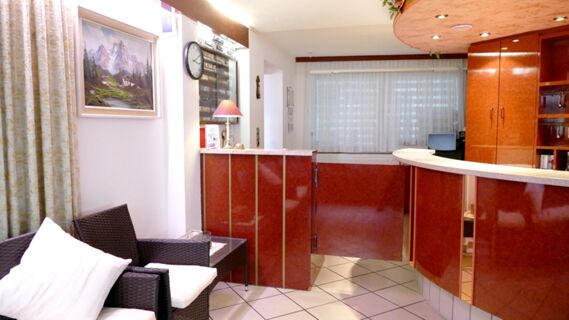Reception area with wicker chair, table, wall clock, mountain landscape painting, and red wood-paneled counters at Hotel & Metzgerei See
