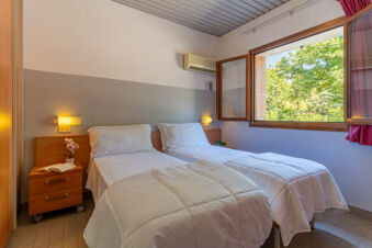 Two single beds with white bedding and pillows under a split air conditioner near an open window showing greenery at Bologna Easy Camping Village
