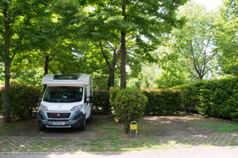 White motorhome parked in a shaded, leafy spot with green bushes and a yellow E 14 sign at Bologna Easy Camping Village