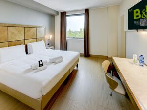 Hotel room at B&B Hotel Padova with two beds, wooden headboard, window with curtains, wall-mounted TV, desk with chair and water bottles